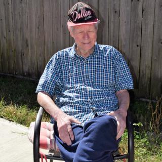 A man wearing a hat and checkered shirt sits outdoors on a chair by a fence.