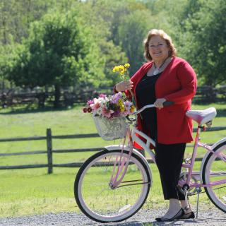 A woman stands beside a bicycle with a basket full of flowers in a scenic outdoor setting.