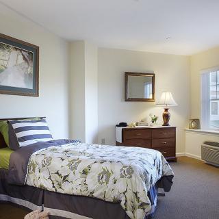 A cozy bedroom with a single bed, decorative pillows, and a dresser topped with a lamp.