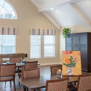 A cozy dining room with tables and chairs, featuring a colorful painting and large windows letting in natural light.