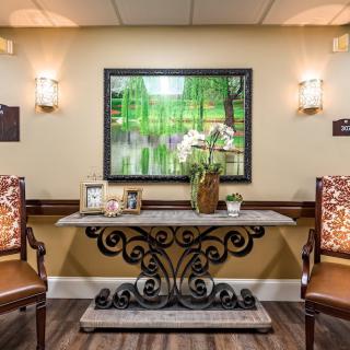 A warmly lit lobby area featuring a decorative table with two chairs, framed artwork, and plants.