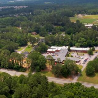 Aerial view of buildings surrounded by lush green trees and a road in a countryside setting.