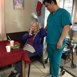 An elderly woman is seated at a table talking on the phone, while a caregiver in scrubs stands nearby in a nursing home setting.