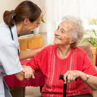 A caregiver smiling at an elderly woman who is sitting with a cane.