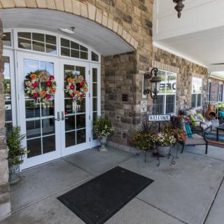 A welcoming entrance with double doors adorned with colorful wreaths and outdoor seating surrounded by decorative plants.