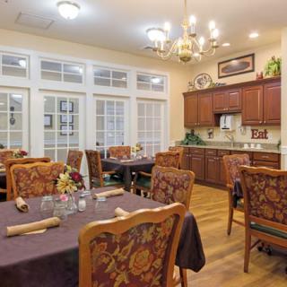 A cozy dining area with neatly arranged tables and upholstered chairs, featuring warm lighting from chandeliers.