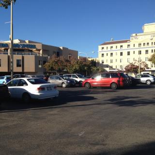 A busy parking lot filled with cars under a clear blue sky, surrounded by urban buildings.