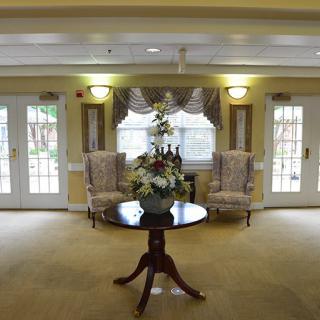 A cozy lobby area with a central table adorned with flowers, flanked by two chairs and surrounded by windows.