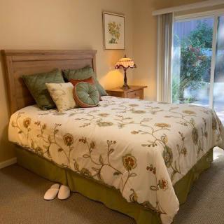 A cozy bedroom featuring a floral comforter on the bed, accent pillows, a bedside lamp on the nightstand, and a pair of slippers on the floor.