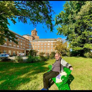 A person sitting on a green bench in a park with a large building in the background on a sunny day.
