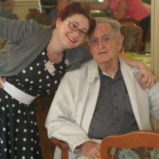 A woman smiling and posing with an elderly man seated indoors.