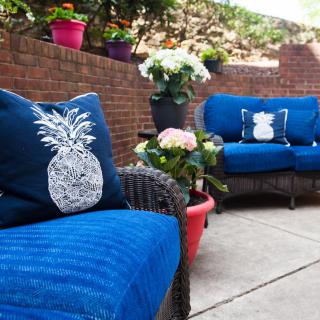 A cozy outdoor patio with wicker furniture featuring blue cushions and pineapple-themed decorative pillows, surrounded by potted plants against a brick wall.