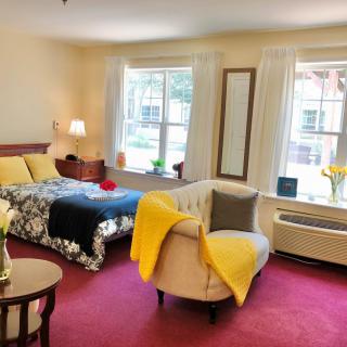 A cozy bedroom with a bed, chair, and large windows, featuring warm decor and red carpet.