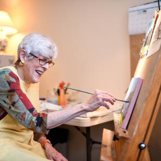 An elderly woman joyfully paints on an easel in her studio.