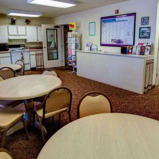 A community room with a kitchen and dining area featuring round tables and chairs on a carpeted floor.