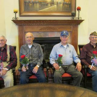 A group of elderly veterans holding roses and an American flag seated in front of a fireplace.