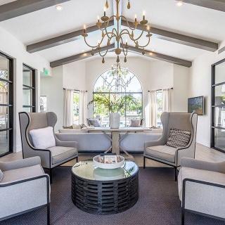 A modern living room with elegant furniture and a central chandelier, featuring ample natural light.