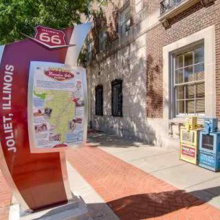 A Route 66 informational sign in Joliet, Illinois, displayed on a sidewalk next to a building.