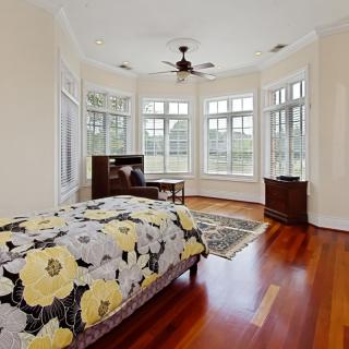 A spacious bedroom with wooden floors, large windows allowing natural light, and a floral bedspread.