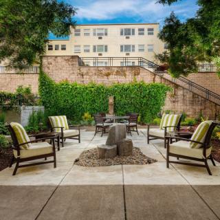 A cozy outdoor patio with chairs surrounding a fire pit, nestled in a courtyard with greenery and a building in the background.