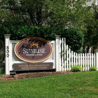 A welcoming sign for Sunrise Retirement, surrounded by greenery and a white picket fence.