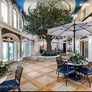 Luxurious indoor atrium featuring a large tree, elegant seating area with tables and umbrellas, surrounded by natural light.