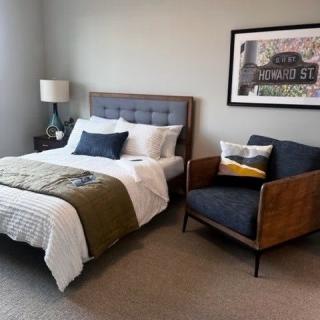 A modern bedroom featuring a cozy bed with white and olive bedding, a stylish armchair, and framed artwork on the wall.