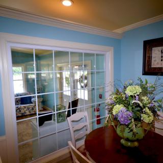 A cozy dining room with blue walls and glass doors opens to a bright living area, featuring a table set with a floral arrangement.