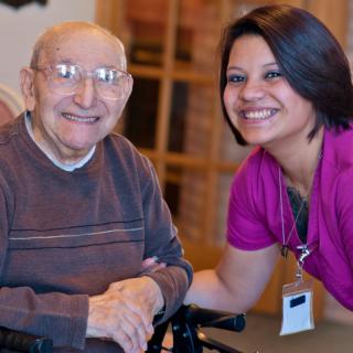 An elderly man in a wheelchair smiling alongside a caregiver indoors.