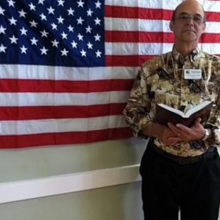 A man stands indoors holding a book in front of an American flag.
