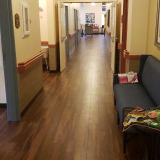 A long hallway with wooden flooring and seating along the side, resembling a healthcare facility.