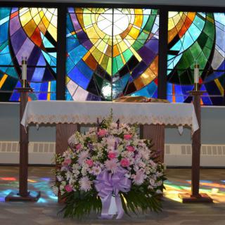 A church altar adorned with floral arrangements, set against an illuminated stained glass backdrop.