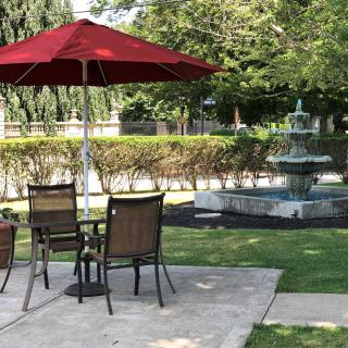 A serene outdoor patio area with a red umbrella, chairs, and a nearby fountain surrounded by greenery.