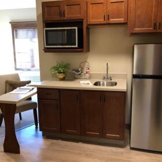 A compact kitchenette with wooden cabinetry, featuring a microwave, stainless steel fridge, and sink.