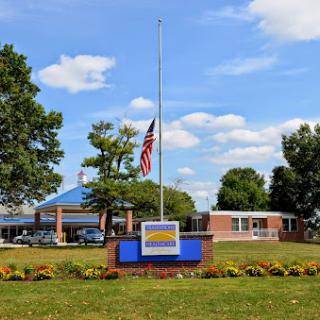 A school building with a flag at half-mast, surrounded by greenery and a clear sky.