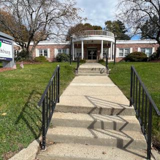 A charming building with a circular porch, surrounded by a well-kept lawn and trees, featuring a paved pathway leading to its entrance.