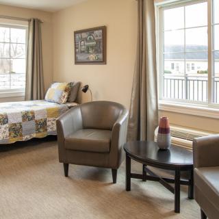 A cozy room with two armchairs, a small table, a bed, and large windows.