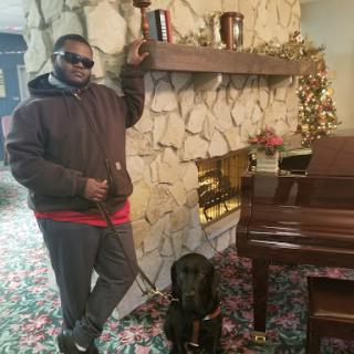 A man poses confidently beside a black labrador dog in a cozy indoor setting decorated for the holidays.