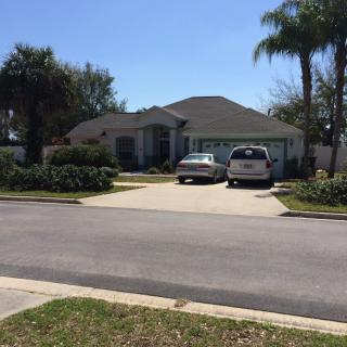 A suburban house with a driveway and two cars parked in front, surrounded by palm trees on a sunny day.