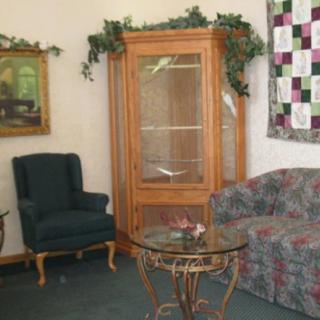 A cozy living room with an armchair, couch, glass table, and a wooden curio cabinet. Decorative elements include a painting on the wall and a quilt.