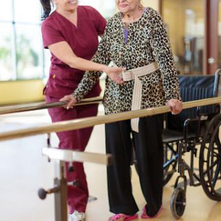 A nurse assists an elderly patient in a rehabilitation setting, supporting her as she practices walking using a rail.