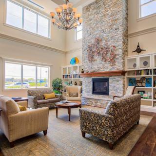 A spacious living room with high ceilings features a central stone fireplace, surrounded by cozy chairs and a large chandelier above, complemented by bookshelves and abundant natural light from the windows.