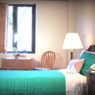 A cozy bedroom featuring a teal bedspread, a bedside lamp, and a window allowing natural light to fill the room.