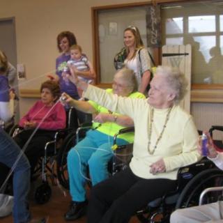 A group of elderly individuals, some in wheelchairs, enjoying a community activity indoors with family and friends.