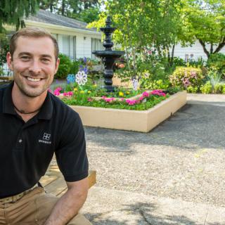 A man smiling in a well-maintained garden with a fountain and blooming flowers.