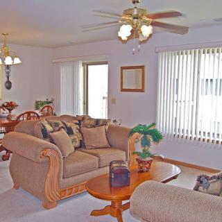 A cozy living room with a matching sofa set, wooden coffee table, and dining area in the background, featuring a ceiling fan and large windows with blinds.