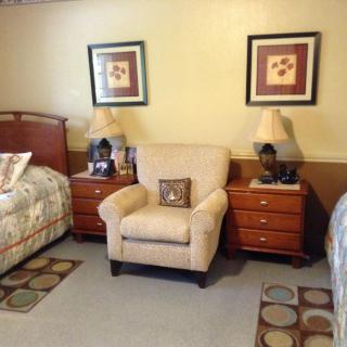 A cozy bedroom with twin beds and an armchair between two nightstands, decorated with lamps and framed pictures.