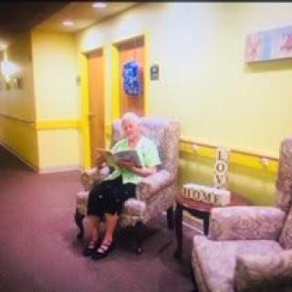 An elderly woman sits in a hallway reading a magazine, surrounded by homey decor.