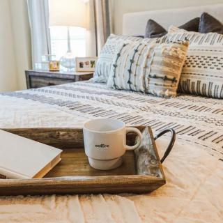 A cozy bedroom with a neatly made bed, featuring decorative pillows and a wooden tray with a cup and notebook.