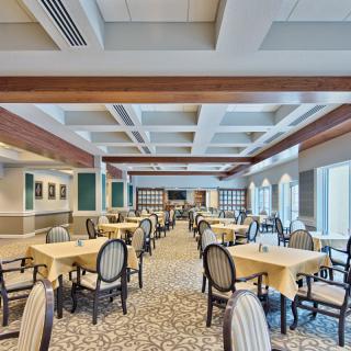 A spacious and elegantly furnished dining area with rows of tables and chairs, featuring large windows and decorative ceiling beams.